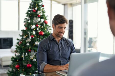 Happy young businessman working on laptop during christmas holiday. Successful business man in modern office working with xmas tree in background. Entrepreneur using laptop in modern office.の写真素材