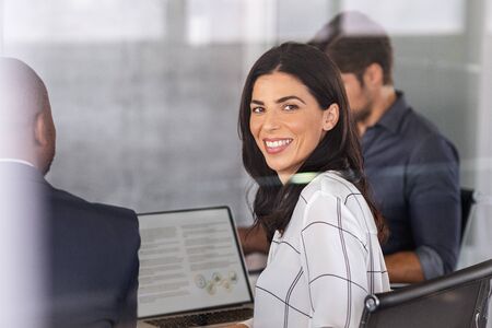 Smiling business woman sitting in conference room during presentation and looking at camera. Latin business woman sitting in board room looking behind window glass. Mature woman in meeting room during brainstorming with associates.の写真素材