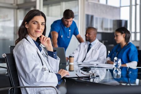 Portrait of mature female doctor sitting in meeting room with specialist and nurses discussing case in background. Successful woman doctor in labcoat and stethoscope in hospital. Smiling pediatrician looking at camera with medical staff brainstorming in background.の写真素材