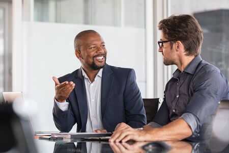 Two happy business colleagues at meeting in modern office interior. Successful african boss in a conversation with young employee in boardroom. Marketing team of two businessmen discussing strategy in meeting room.の写真素材