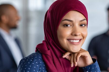 Portrait of young muslim woman wearing hijab in office while looking at camera. Close up face of arabic business woman covered with headscarf smiling. Successful arab businesswoman in modern office.の写真素材