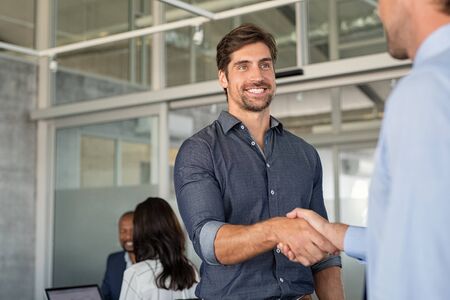 Two cheerful businessmen shaking hands after an agreement. Two confident business men shaking hands and smiling while standing in modern office with copy space. Young smiling business man making handshake for greeting and dealing.の写真素材