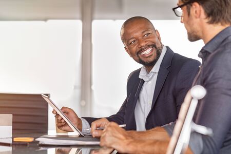Business partners working on project together. Smiling african businessman working and discussing with employee in modern office over a report. Happy and successful leader with colleague in conference room discussing new project.の写真素材