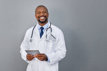 African american doctor holding digital tablet against grey background. Smiling mature doctor in labcoat standing against gray background. Successful happy healthcare worker looking at camera, copy space.の写真素材