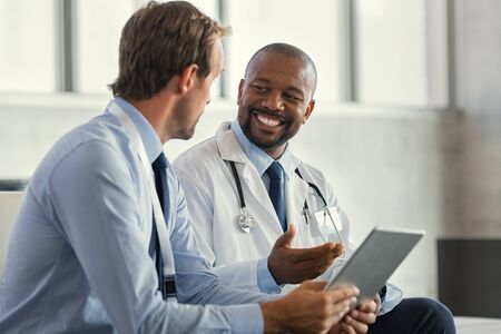 Two mature smiling doctors having discussion about patient diagnosis, holding digital tablet. Successful african surgeon discussing case after positive result. Representative pharmaceutical discussing with african happy doctor about new medicine.の写真素材