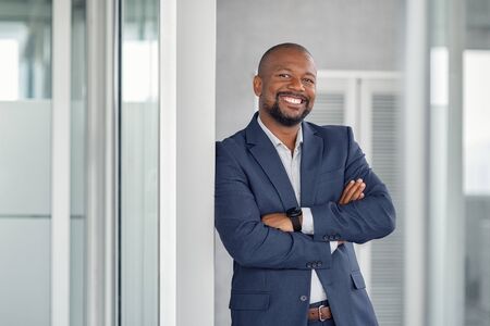 Mature cheerful african american executive businessman at workspace office. Portrait of smiling ceo at modern office workplace in suit looking at camera. Happy leader standing in front of company building.の写真素材