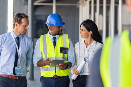 Team of construction workers discussing project details with executive supervisor holding blueprints and digital tablet. Team of architects and civil engineers inspecting construction site. Structural african engineer and architect with work vests discuss the construction process.の写真素材