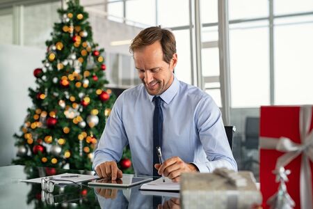 Mature business man working with digital tablet with xmas present and gift on desk. Successful formal businessman using digital tablet to send an email while sitting in modern office. Happy entrepreneur working in office during christams holiday.の写真素材