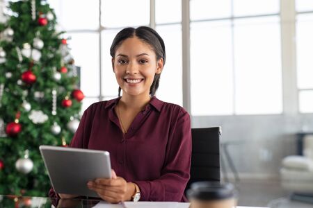 Happy business woman using digital tablet in modern office with christmas tree in background. Young middle eastern woman smiling and looking at camera during xmas. Portrait of cheerful young businesswoman working during christmas time with copy space.の写真素材