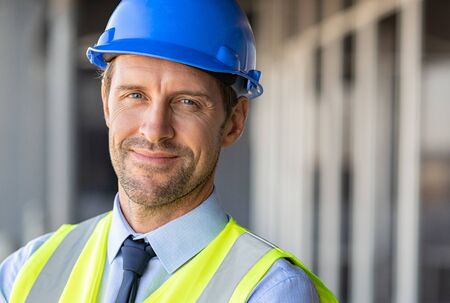 Successful male architect at building site looking at camera. Confident construction manager wearing blue helmet and yellow safety vest with copy space. Portrait of successful mature civil engineer.の写真素材