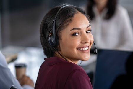 Portrait of smiling young businesswoman wearing headset in office. Portrait of a customer service agent looking at camera. Happy telephone operator latin girl with headset working at computer.の写真素材