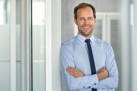 Portrait of a successful leader smiling at modern office with copy space. Happy executive businessman looking at camera at work place. Mature entrepreneur standing with crossed arms leaning on glass wall with copy space.の写真素材