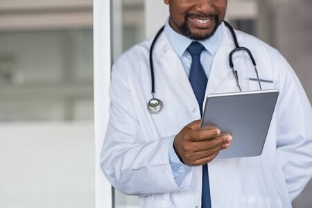 Mature black doctor using digital tablet at hospital.Â Close up of smiling doctor at private clinic reading medical exam on tablet. Happy practitioner holding digital report for patient's health record in medical clinic.の写真素材