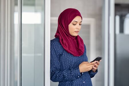 Young muslim businesswoman leaning against glass wall using smartphone. Islamic business woman in hijab using mobile phone in modern office. Arabic serious girl texting on cellphone to send email message.の写真素材