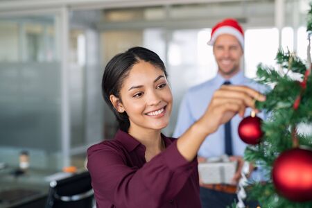 Portrait of smiling latin business woman decorating christmas tree in office. Happy middle eastern woman decorating tree for christmas holiday. Multiethnic people celebrating xmas holiday at work.の写真素材