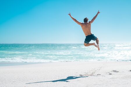 Back view of happy young man jumping with outstretched arms on the beach with copy space. Rear view of shirtless guy wearing swimwear and having fun on tropical beach during vacation. Carefree and freedom concept.の写真素材