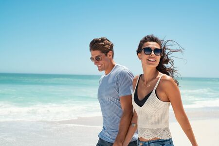 Beautiful woman with man wearing sunglasses walking on beach.. Young couple enjoying honeymoon after marriage at sea. Happy casual couple holding hands and walking at the beach with copy space.の写真素材
