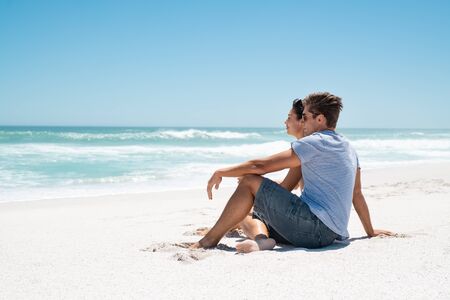 Young couple sitting on sand by sea looking at the horizon and thinking about the future. Happy man and beautiful woman enjoying romantic vacation at sea. Thoughtful couple relaxing on beach and contemplating the ocean, copy space.の写真素材