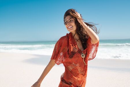 Beautiful latin woman running on tropical beach with copy space. Happy glamour and carefree woman walking on beach while looking behind. Stylish girl in red lace dress with black bikini enjoying summer vacation at sea.の写真素材