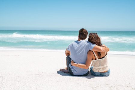 Rear view of young couple sitting on tropical beach and embracing while looking the ocean. Thoughtful man embracing loving woman on the beach while contemplating the horizon with copy space. Back view of romantic couple sitting together on beach and thinking about the future.の写真素材