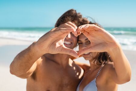 Happy husband and wife gesturing heart with fingers at sea during honeymoon. Young couple showing heart shape with hand gesture. Smiling couple looking through a heart shape made with their fingers at beach.の写真素材