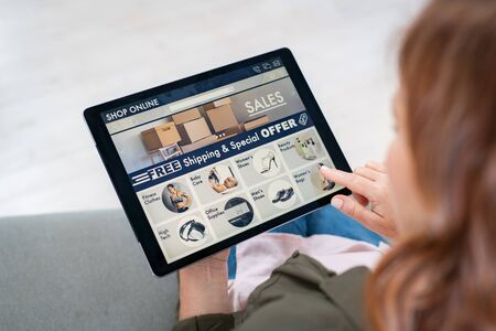 Back view of mature woman doing shopping online on website with digital tablet at home. Top view of woman with red hair relaxing on sofa while shopping online on ecommerce website. Rear view of girl hand touching screen while selecting product on e-commerce portal.の写真素材