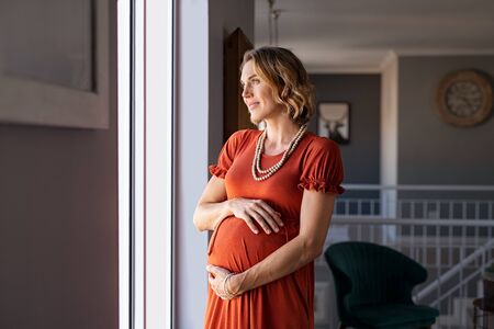 Portrait of mature pregnant woman at home looking outside the window during maternity. Thoughtful pregnant woman standing near window and thinking about the future family. Smiling lady in gestation caressing her baby bump with pensive look.の写真素材