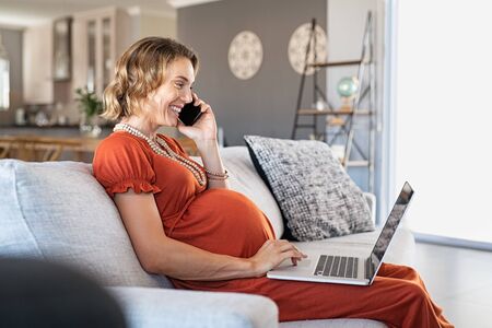 Pregnant mid adult woman using laptop and talking on mobile phone. Happy expectant businesswoman sitting on couch at home in smart working while speaking on smartphone. Smiling woman in maternity working on computer and talking with a client on cellphone.の写真素材