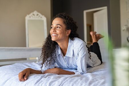 Happy african american woman lying on front and thinking on bed. Thoughtful mixed race girl lying on bed while looking up in contemplation. Smiling young latin woman daydreaming about future while relaxing at home.の写真素材