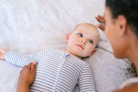Portrait of toddler lying on bed with her mother. Close up face of cute relaxed infant looking at mother playing with copy space. Rear view of multiethnic woman playing with adopted child lying on bed.の写真素材