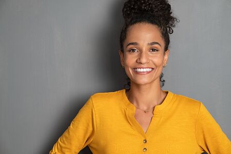 Portrait of happy young brazilian woman standing on gray wall and looking at camera. Confident african american girl isolated against grey background with copy space. Cheerful beautiful mixed race woman with curly hair smiling.の写真素材