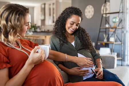 Smiling woman with hands on expecting mother's stomach feeling baby movement while sitting on couch at home. Lesbian couple both with a baby bump relaxing at home. Happy friend feeling belly of young african pregnant woman.の写真素材