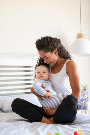 Portrait of loving young latin woman bonding with her newborn baby, sitting on bed at home. Happy...