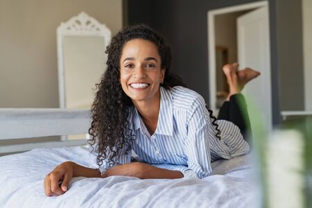 Cheerful young multiethnic woman looking at camera and smiling while lying on bed. Pretty casual mixed race woman lying on front in barefoot on her bedroom. Portrait of beautiful african american girl resting at home.の写真素材
