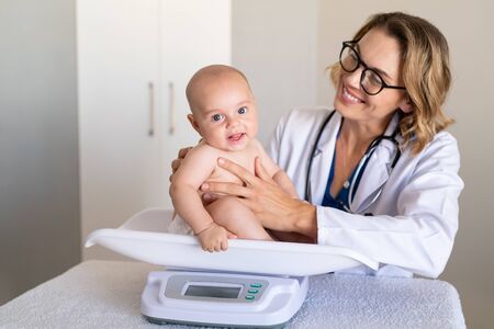 Adorable baby boy sitting on scale during weight measuring exam at doctor office. Pediatrician weighting baby girl on scales. Child specialist making an assessment of a babyâs growth progress.の写真素材