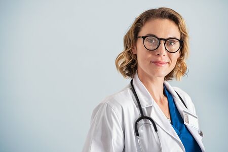 Portrait of happy mid adult smiling doctor looking at camera. Happy beautiful mature pediatrician standing over blue background with copy space. Reliable general practitioner woman with stethoscope and labcoat isolated over background.の写真素材