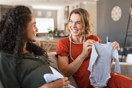 Happy pregnant woman looking at baby clothes with joyful african friend. Laughing pregnant friends sitting on sofa and looking at baby shower gifts. Excited two expecting girls checking baby clothes for new baby.の写真素材