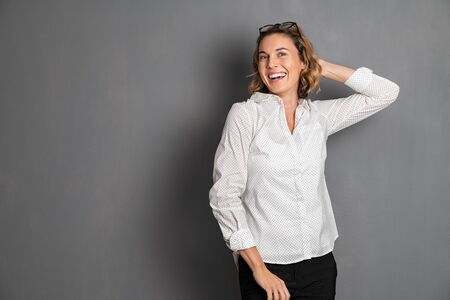 Portrait of excited middle aged woman smiling against grey background and looking at camera. Cheerful young businesswoman in formal relaxing after work. Beautiful german girl posing and dancing for camera with big grin and copy space.の写真素材