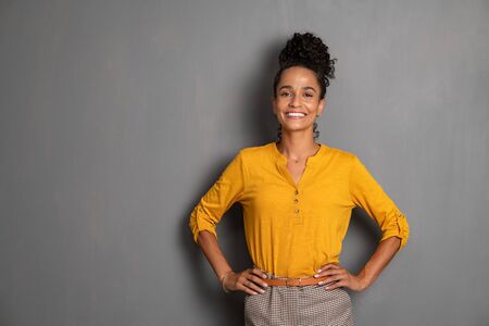 Portrait of young african woman standing with hands on waist and looking at camera. Confident stylish latin girl smiling isolated against grey background. Beautiful cool woman with copy space standing on gray wall.の写真素材