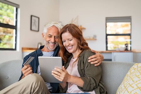 Happy mature couple sitting on sofa and doing online shopping on digital tablet at home. Senior husband and smiling wife paying bills online. Joyful middle aged couple doing online payment on digital tablet, copy space.の写真素材