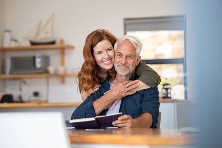 Portrait of happy mature couple at home looking at camera. Romantic wife embracing senior husband from behind while laughing together. Portrait of beautiful woman in love hugging  old man in perfect harmony.の写真素材