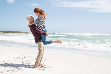 Mature couple hugging on the beach on a sunny day with copy space. Happy wife hugging her husband at the beach with her feet off the ground. Loving senior man lifting beautiful woman above the ground hugging her on the beach with sea in the background.の写真素材