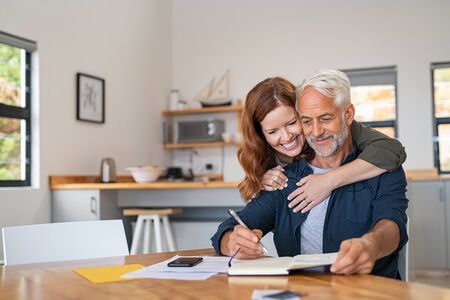 Loving mature wife embracing husband from behind while writing in book. Happy middle aged couple making to do list of purchases and discussing future plans. Cheerful senior man working at home on wooden table with beautiful woman hugging him from behind, copy space.の写真素材