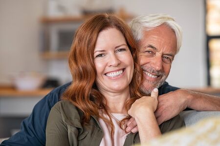 Portrait of middle aged couple hugging and looking at camera. Close up face of happy mature couple having fun at home. Senior man embracing his beautiful wife on the couch while looking at camera together.の写真素材