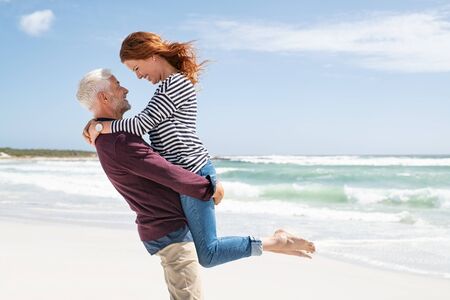 Mature couple hugging on the beach on a sunny day. Senior active man lifting beautiful mid woman on the beach with sea in the background and copy space. Happy retired couple hugging at seaside while looking at each other.の写真素材