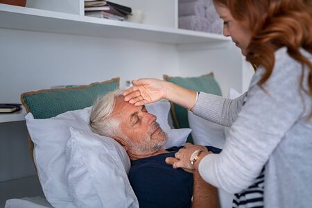 Mature woman checking fever temperature of senior man lying on bed with closed eyes. Old husband resting at home feeling sick while wife measure the fever by touching forehead. Elderly man feeling the seasonal symptoms of the flu.の写真素材