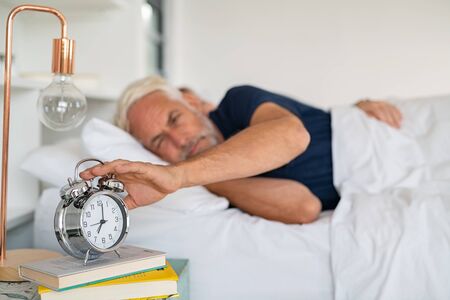 Old sleepy man lying on bed and stopping alarm clock with his wife in background. Senior man lying on side on bed turning off an alarm clock in the morning at 7am. Mature adult waking up in the morning.の写真素材