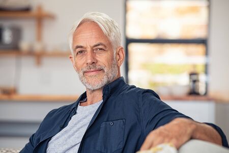 Handsome senior man with white hair sitting on couch and looking at camera. Casual satisfied old man relaxing on sofa while looking at camera. Portrait of proud mature man stay at home with copy space.の写真素材