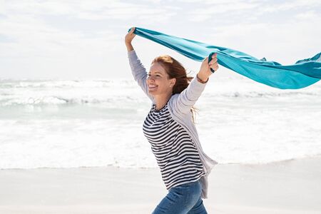 Happy smiling woman running with lightblue scarf enjoying at beach. Beautiful redhead woman holding blue fabric at wind in a summer vacation. Middle aged mature woman having fun at seashore with copy space: freedom and carefree concept.の写真素材
