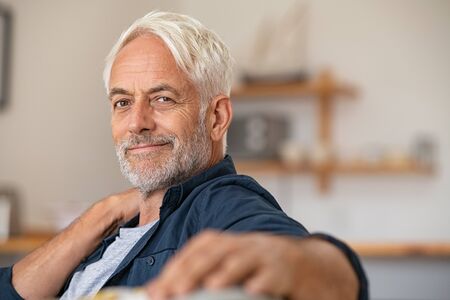 Portrait of a smiling mature man relaxing on couch with copy space. Confident senior man resting at home and looking at camera. Satisfied retired man with grey hair sitting on sofa at home.の写真素材